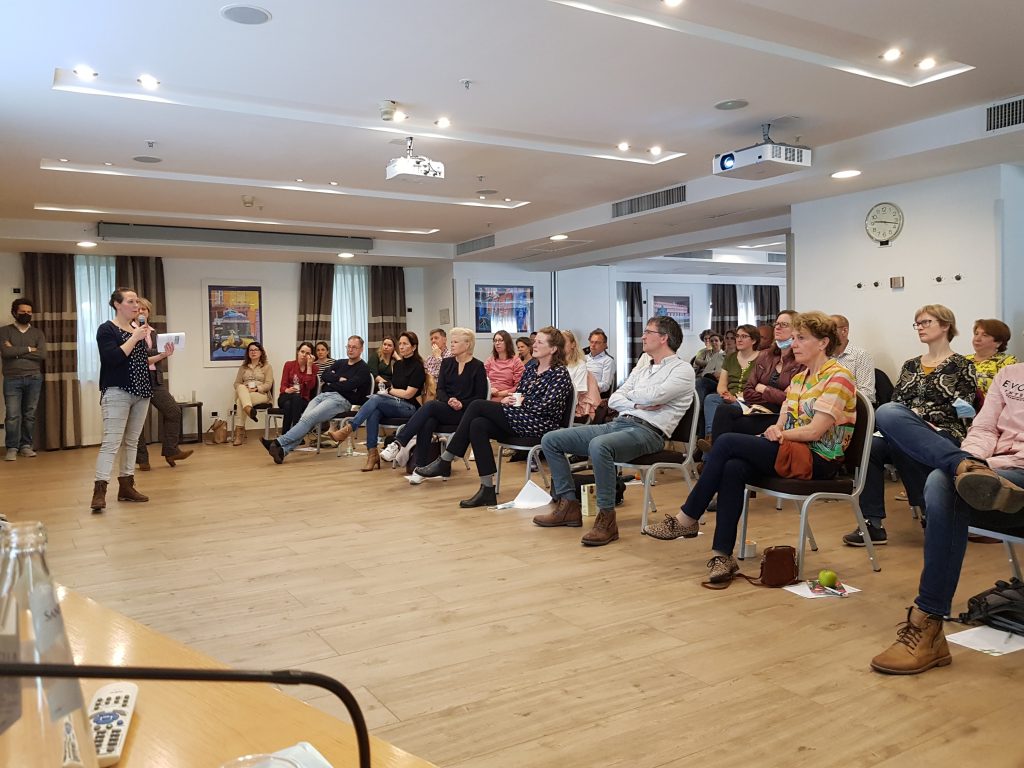 Groep mensen zit op stoelen in een verlichte zaal en luistert naar een vrouw tijdens OverEINDse dagen 2023, met projector en klok.