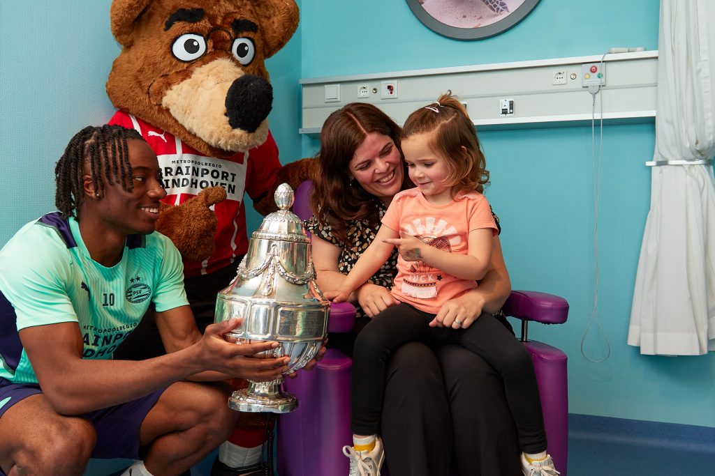 Voetballer bezoekt kinderafdeling Catharina Ziekenhuis, toont trofee aan vrouw en meisje; mascottebeer staat erachter.