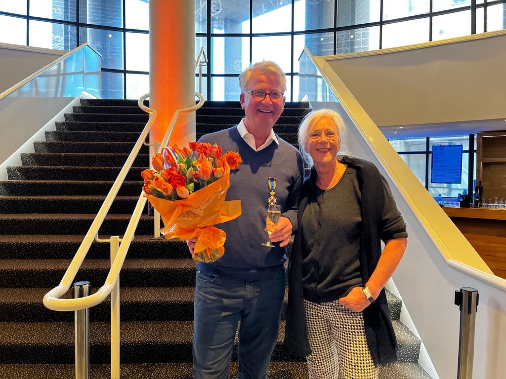 Dick Schoot ontvangt bloemen en een trofee naast een vrouw op een val, ter ere van zijn Ridder in de Orde van de Nederlandse Leeuw.