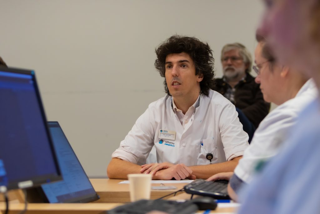 Persoon in witte jas met naamkaartje aan tafel, anderen en computerschermen op achtergrond bij Catharina Ziekenhuis.