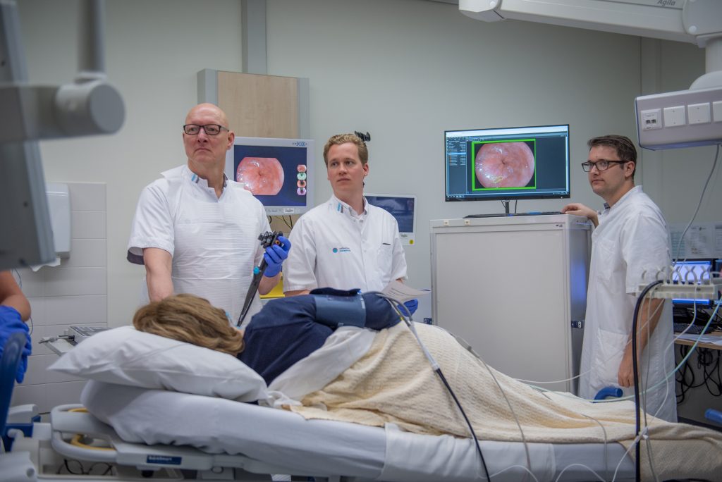 Vier artsen in witte jassen doen een endoscopie bij een patiënt; medische schermen tonen interne beelden in klinische ruimte.
