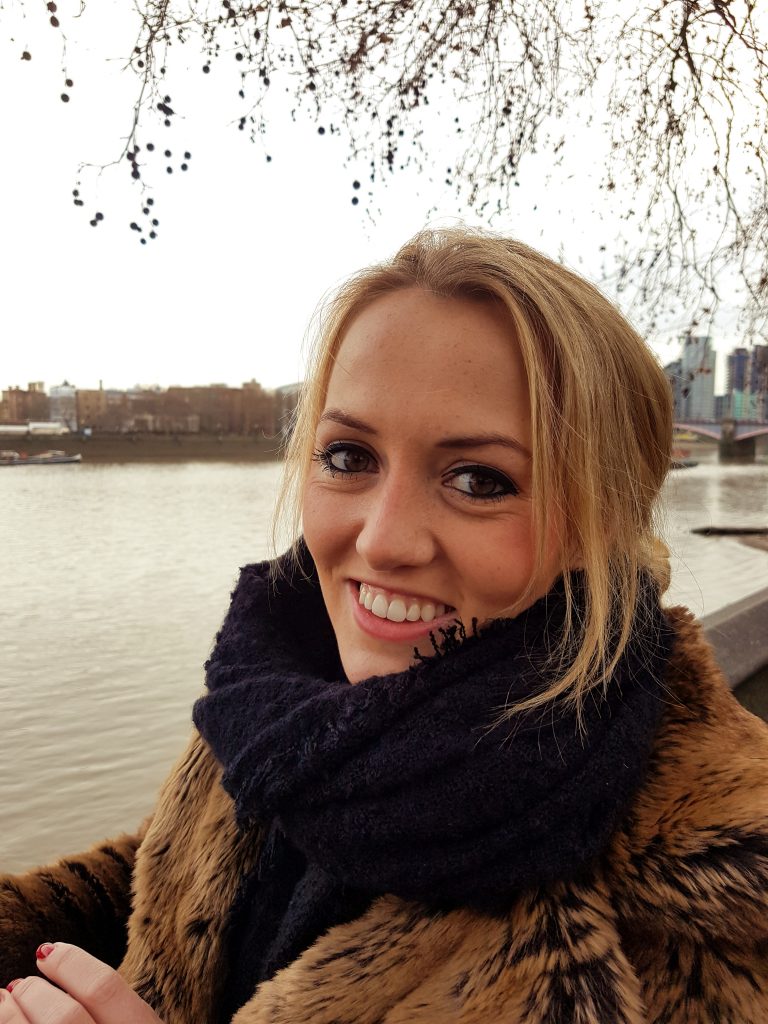 Vrouw met blond haar in bruine bontjas en zwarte sjaal lacht bij rivier; boerenkool takken en gebouwen op de achtergrond.