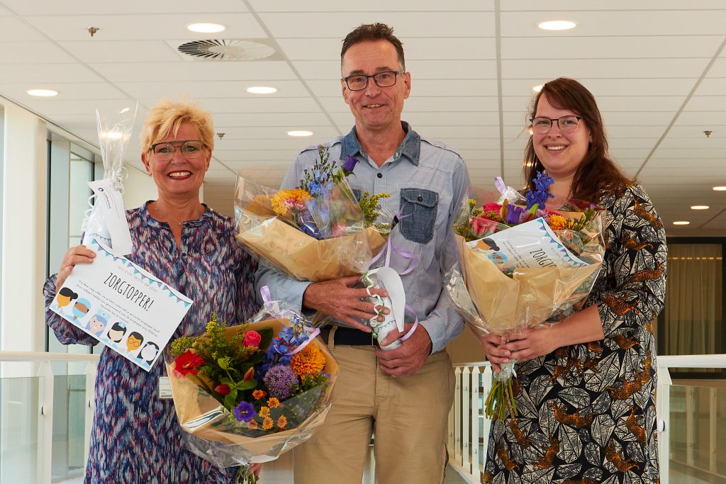Anja, Peter en Sanne staan binnen met boeketten en oorkondes na hun Zorgtoppers 2022 uitreiking. Peter houdt ook een wit object vast.