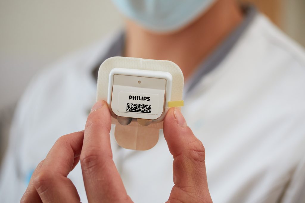 Persoon met witte jas en mondkapje toont PHILIPS medisch sensormet QR-code op huidpleister in Catharina Ziekenhuis. Vage achtergrond.