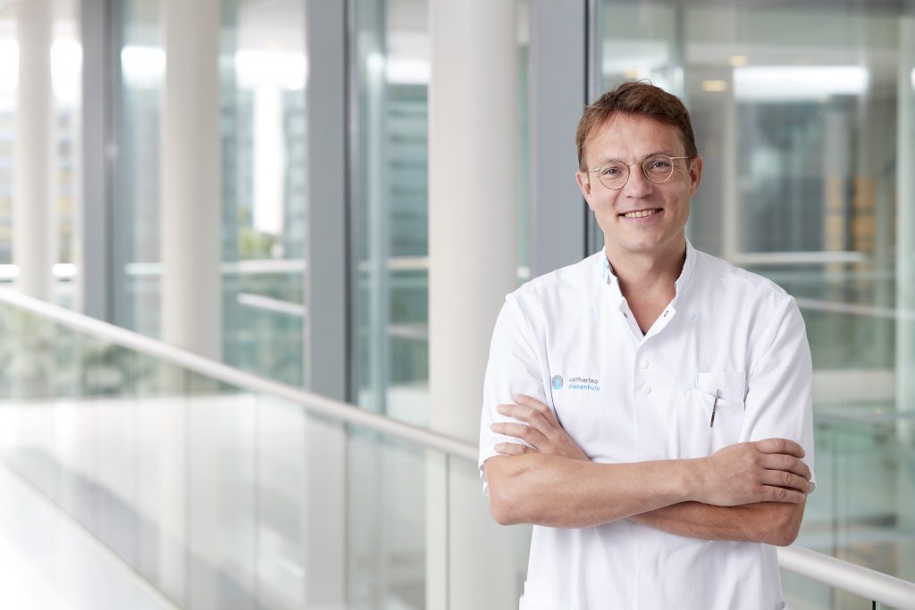 Een persoon in witte doktersjas staat binnen, armen over elkaar en glimlachend. Op de achtergrond moderne glazen ramen.