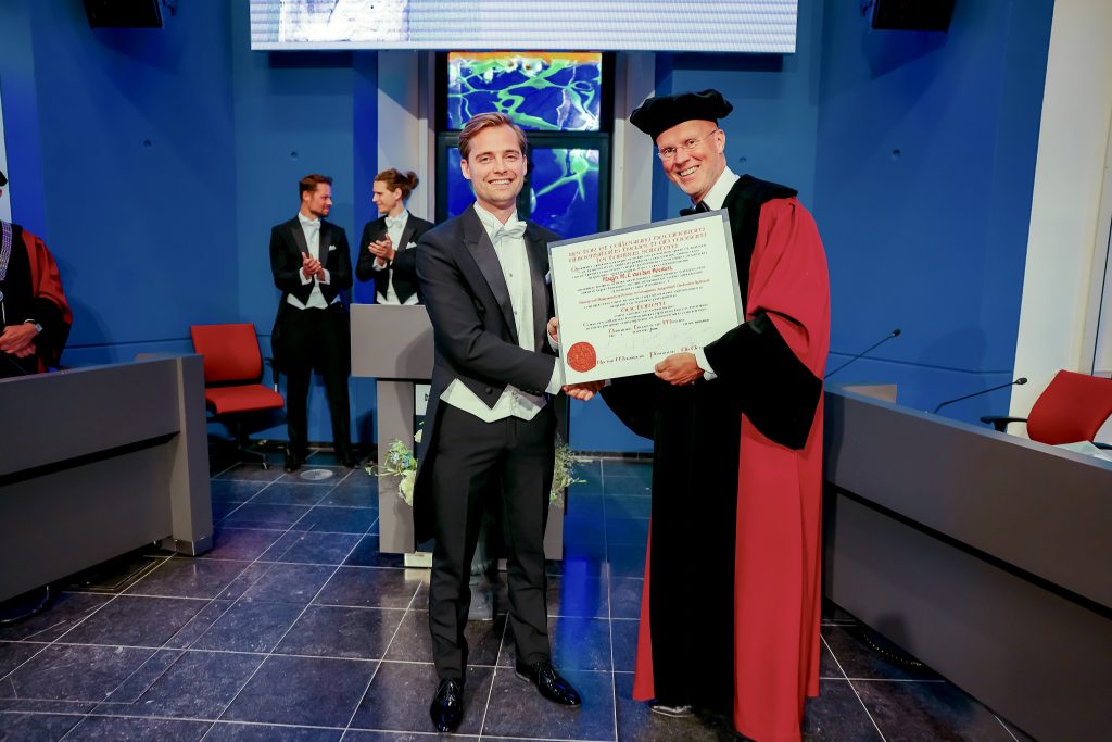 Een man in pak krijgt op het podium een diploma en schudt handen met iemand in toga; drie mensen klappen op de achtergrond.