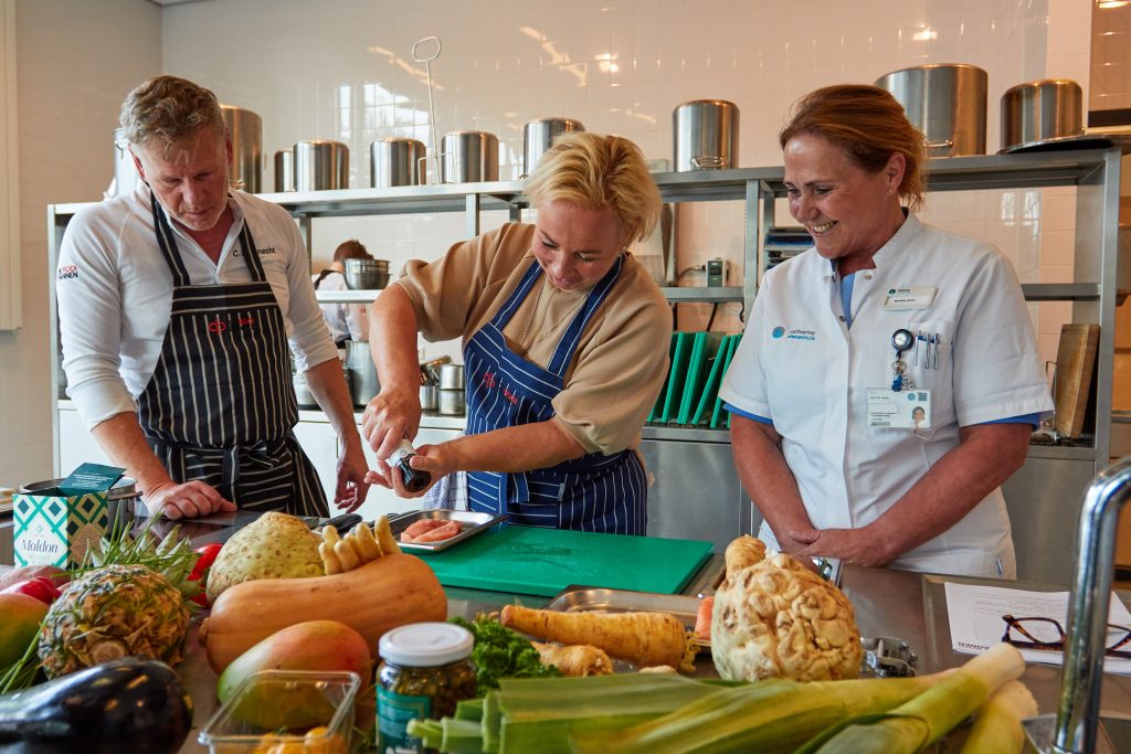 Drie mensen in een keuken bij Rooi Pannen: twee in koksjas, één in medisch uniform. Groenten en fruit op het aanrecht.