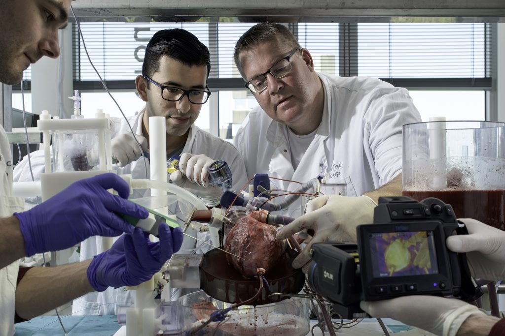 Drie mensen in labjassen werken aan een open hart in het Catharina Ziekenhuis, met slangen, camera en rode vloeistof op tafel.