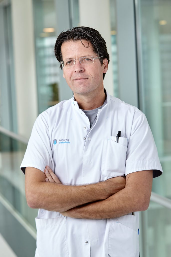 Man met bril en witte doktersjas staat met gekruiste armen in Catharina Ziekenhuis, donkere haren, pennen op zak.