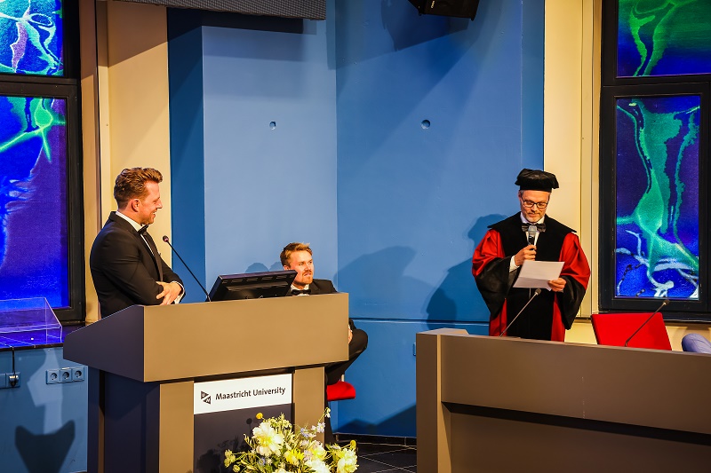 Koen Rovers promoveert aan Maastricht University, met drie mannen bij een podium in een zaal met blauwe muren en glas-in-loodramen.