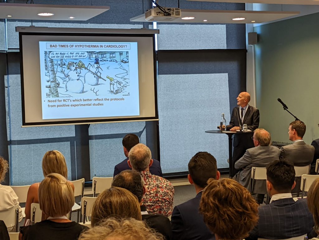 Man in pak presenteert op temperatuursymposium; dia toont cartoon over hypothermie en belang van betere klinische protocollen.