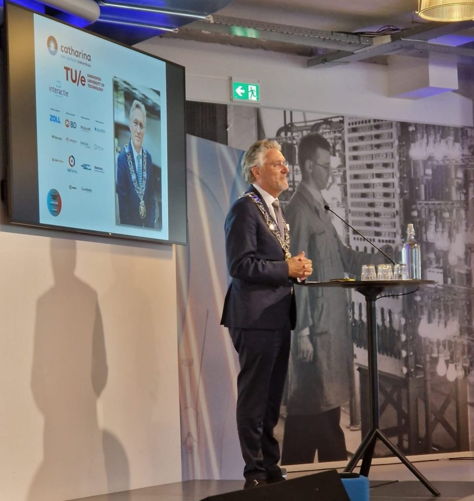 Man in pak met ambtsketen spreekt op het temperatuursymposium; achter hem scherm met foto's, logo's en labfoto.