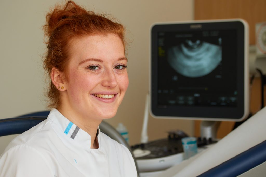 Vrouw in witte medische kleding lacht naar de camera, met echoapparaat en zwart-wit beeld op de achtergrond.