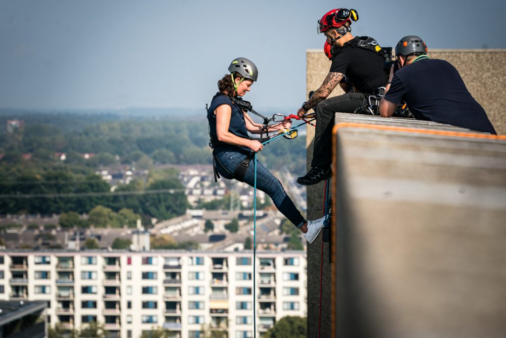 Vrouw in veiligheidsuitrusting daalt met hulp van twee begeleiders langs hoog gebouw bij Catharina Ziekenhuis, stedelijk uitzicht.