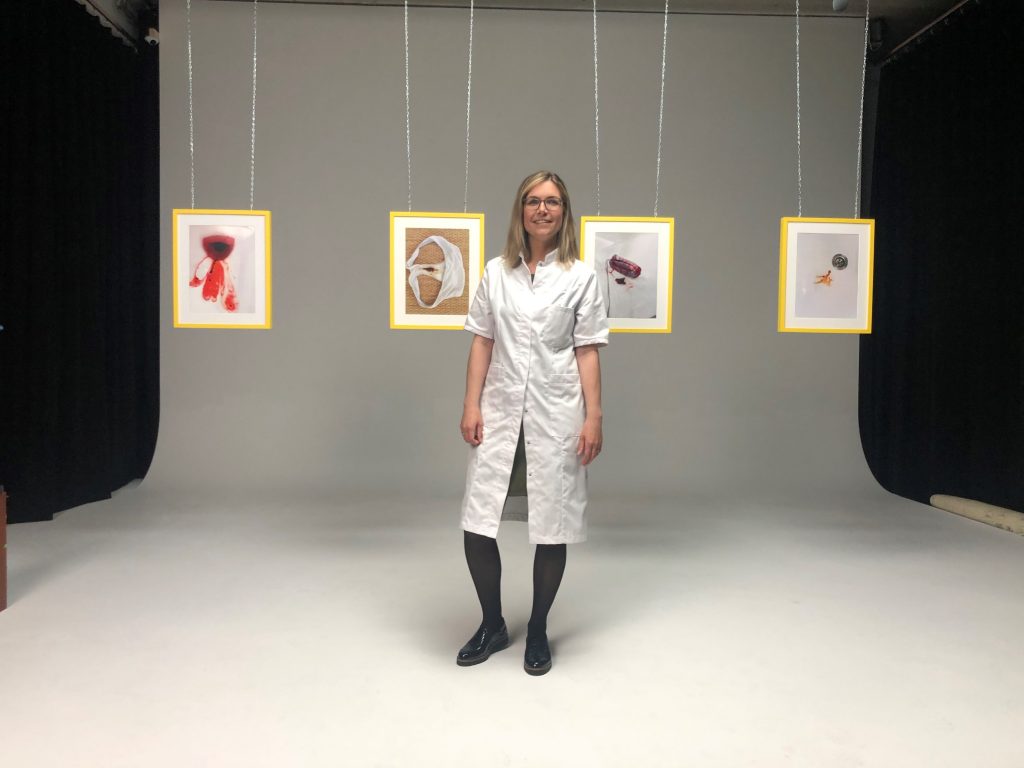 Vrouw in witte jas en zwarte schoenen in studio; vijf kunstwerken met gele randen over sneller herstel na sterilisatie achter haar.