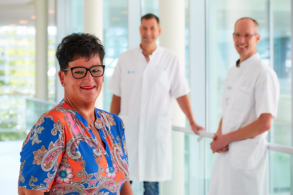Vrouw in kleurrijke blouse glimlacht; twee mannen in witte zorgkleding staan achter haar in modern gebouw met grote ramen.