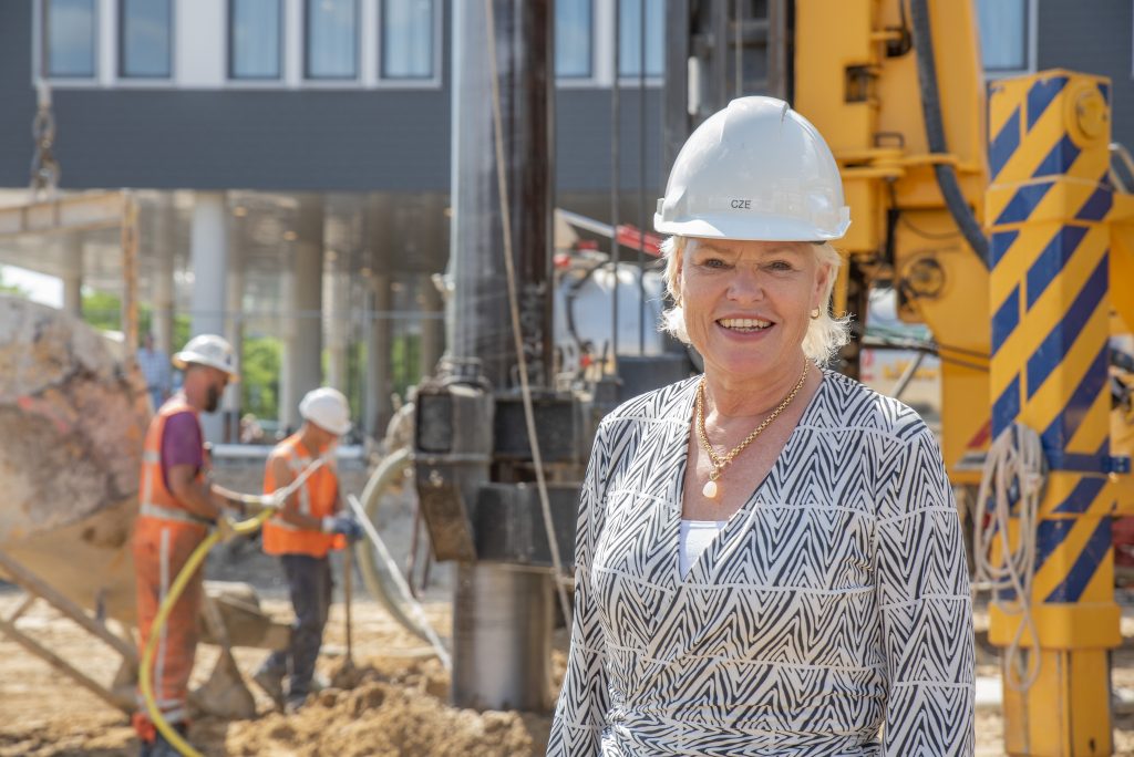 Vrouw met wit bouwhelm en blouse glimlacht op gasloze bouwplaats Catharina Ziekenhuis, met duurzaamheidswerk op de achtergrond.