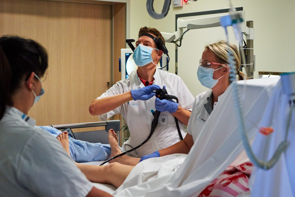 Drie specialisten met maskers en handschoenen voeren een ingreep uit bij een patiënt, met endoscoop en live meekijken.