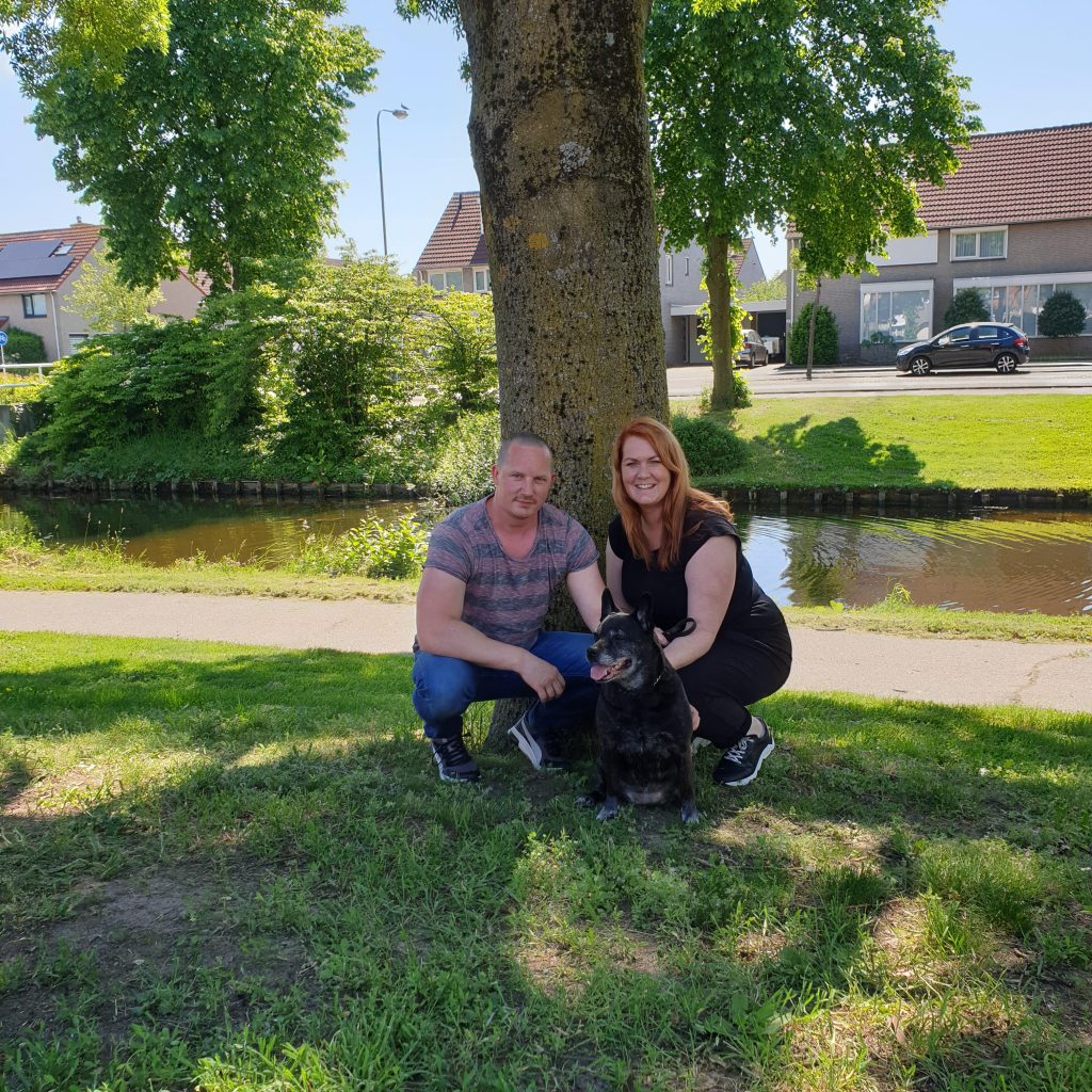 Een man en een vrouw knielen op gras naast een zwarte hond onder een boom, bij een kanaal met huizen op de achtergrond.