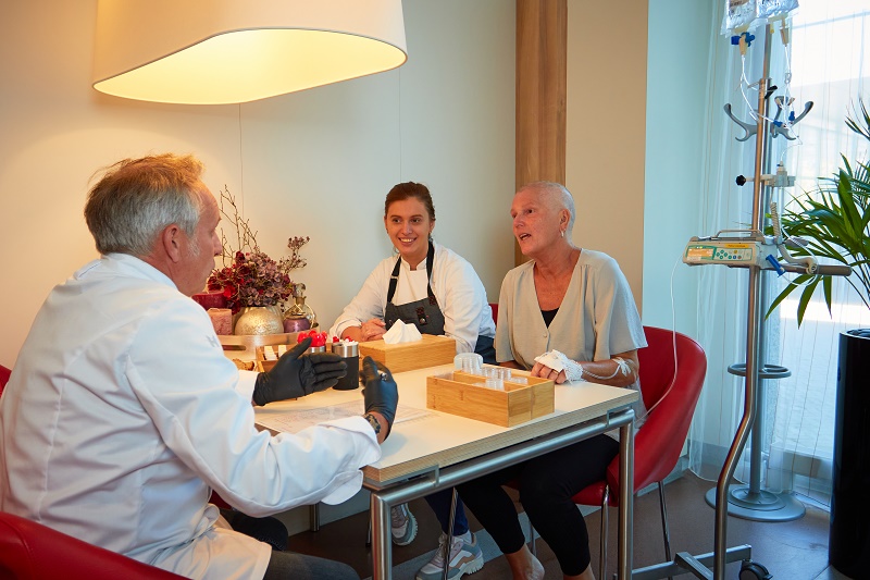 Een zorgverlener en een vrouw met kort haar en polsbandje zitten aan een tafel met medische spullen; iemand kijkt mee.