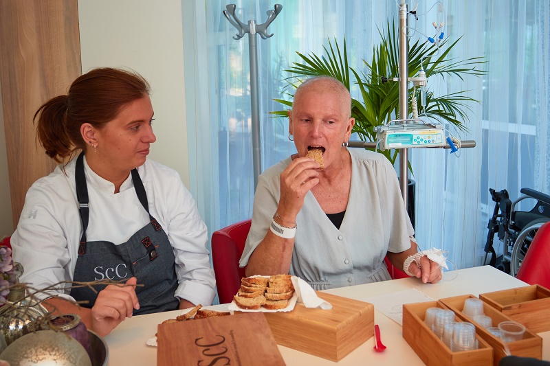 Vrouw in grijs shirt eet brood aan tafel naast chef; op achtergrond brood, dienbladen, bekers en medische apparatuur.