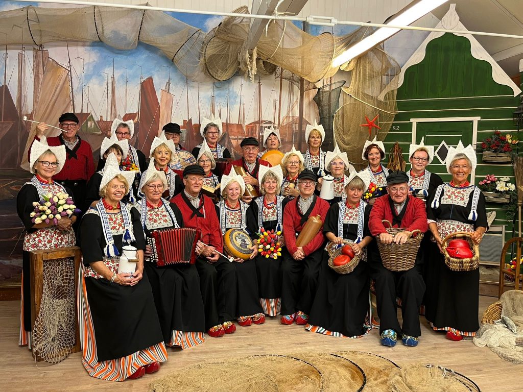 Groep mensen in Volendamse klederdracht, waaronder Catharina vrijwilligers, poseren binnen voor decor met boten en visnetten.