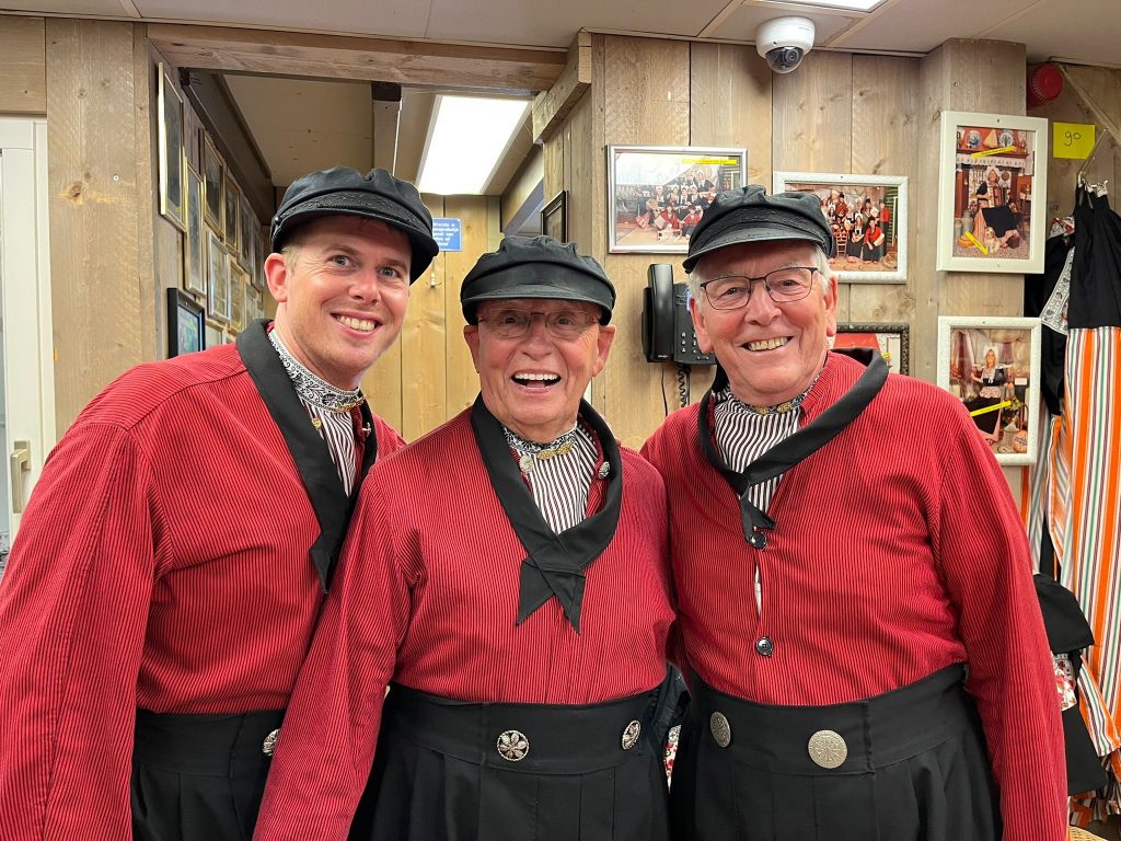 Drie mannen in rode traditionele blouses, zwarte hoeden en dassen poseren binnen bij houten wanden en ingelijste foto's.