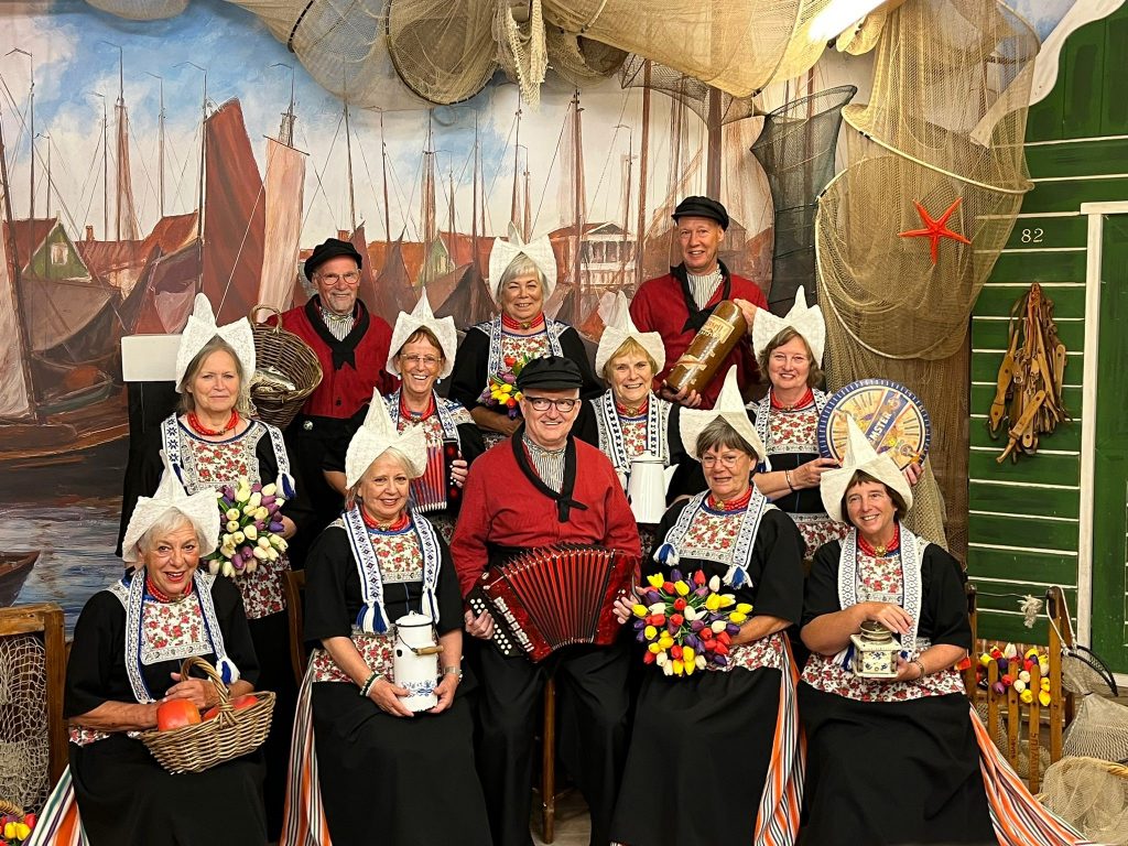 Mannen en vrouwen in Volendammer klederdracht poseren binnen, met Nederlandse attributen en accordeon, voor geschilderde boten.