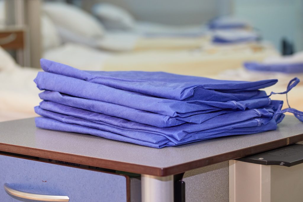 Stapel opgevouwen blauwe OK-jassen op grijze tafel in Catharina Ziekenhuis, ziekenhuisbedden op achtergrond.