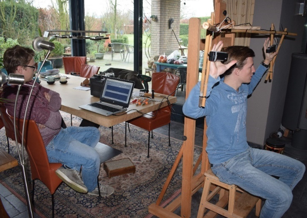 Twee mensen in een ruimte; één werkt met laptop en elektronica voor fitnessapparatuur, de ander onder een houten frame met sensoren.