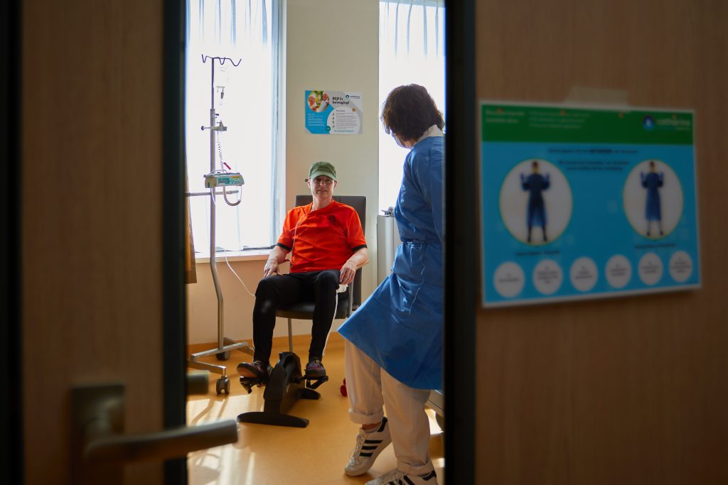 Een persoon met rode trui en huisdier zit in een stoel met infuus; zorgmedewerker in blauwe jas staat ernaast, gezien door deuropening.