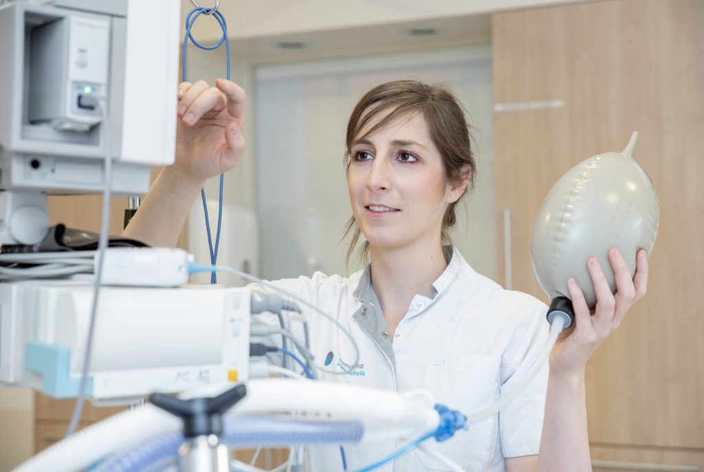Een zorgprofessional in een witte jas stelt een medisch apparaat af en houdt een beademingsballon in een klinische omgeving.