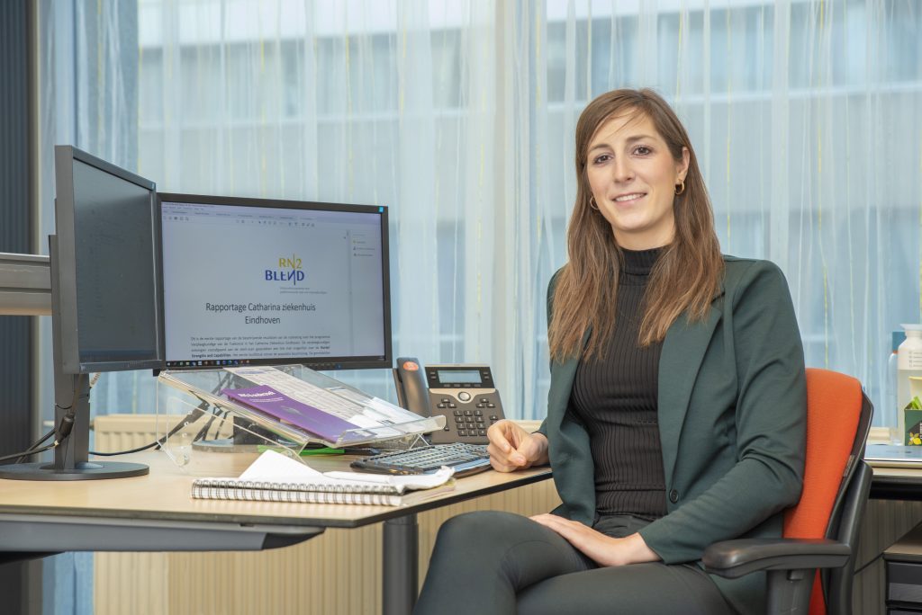 Vrouw aan bureau met twee computerschermen, telefoon en kantoorspullen. Ze lacht naar de camera en bekijkt een rapport.