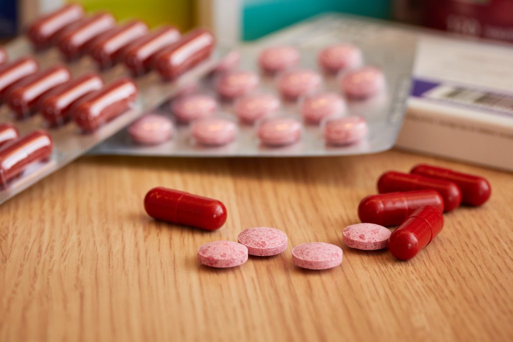Rode capsules en roze tabletten op hout, met daarachter blisterverpakkingen en medicijndoosjes voor gebruik bij vitaminetekort.