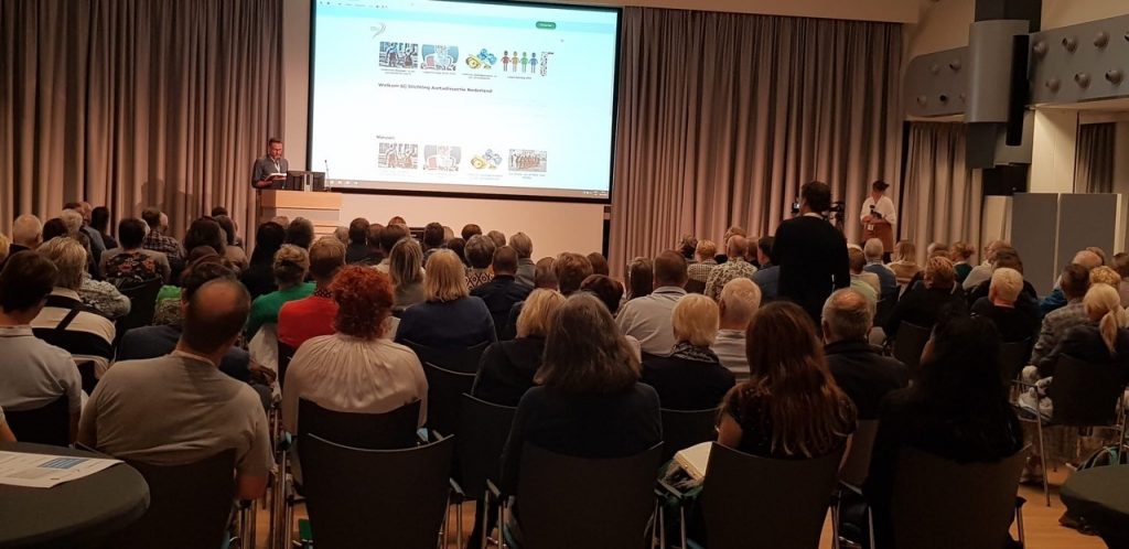 Patiënten zitten in een zaal met grijze gordijnen, kijken naar een spreker met dia's; enkelen staan langs de randen.
