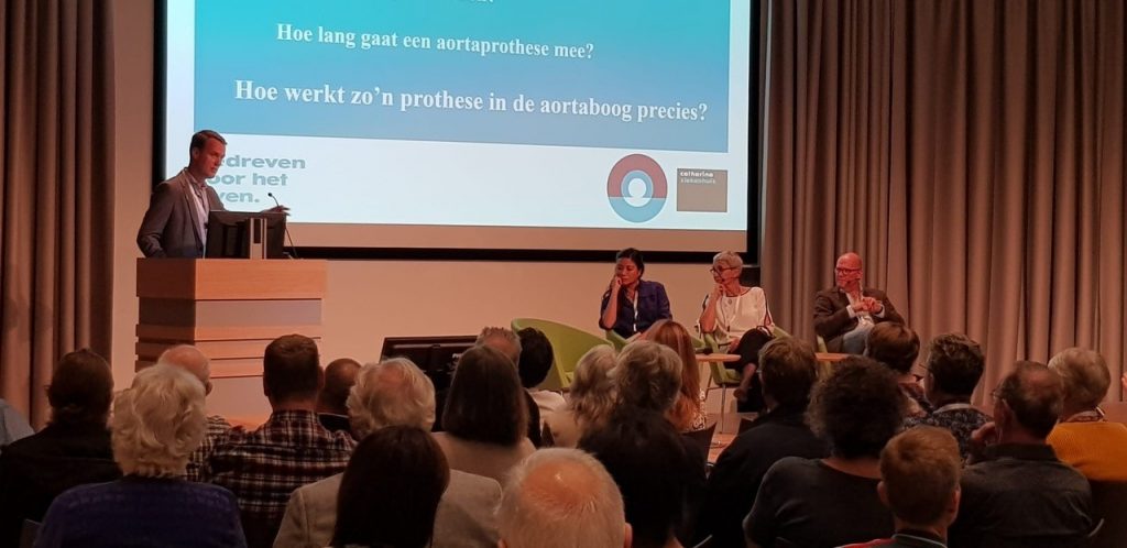 Man spreekt aan een katheder op lotgenotendag; drie mensen zitten erbij, scherm toont medische vragen over aortadissectie.