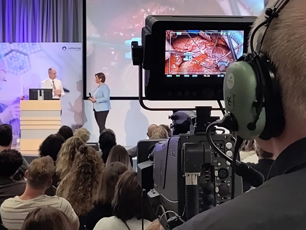 Een cameraman filmt een podiumpresentatie van Omroep Brabant met twee sprekers en een openhartoperatie op het scherm.