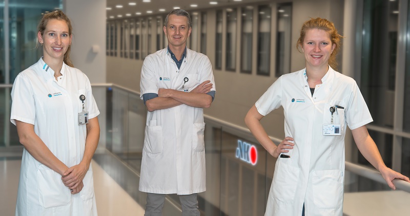Drie artsen in witte jassen staan in een heldere ziekenhuisgang; twee vrouwen vooraan, man erachter, allen kijken naar de camera.