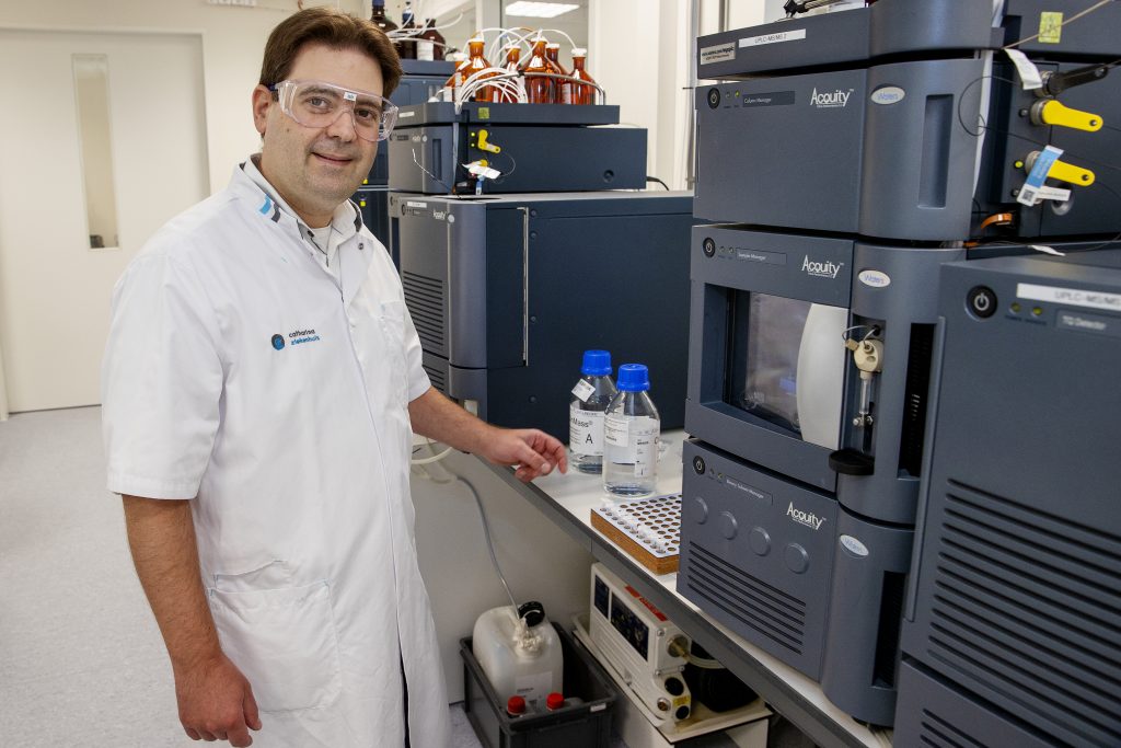 Man in witte labjas en veiligheidsbril in laboratorium naast apparatuur en chemicaliën, kijkt in de camera.