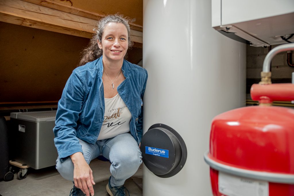 Vrouw met krullen, blauwe jas en spijkerbroek knielt bij witte Buderus-ketel in technische ruimte Catharina Ziekenhuis.