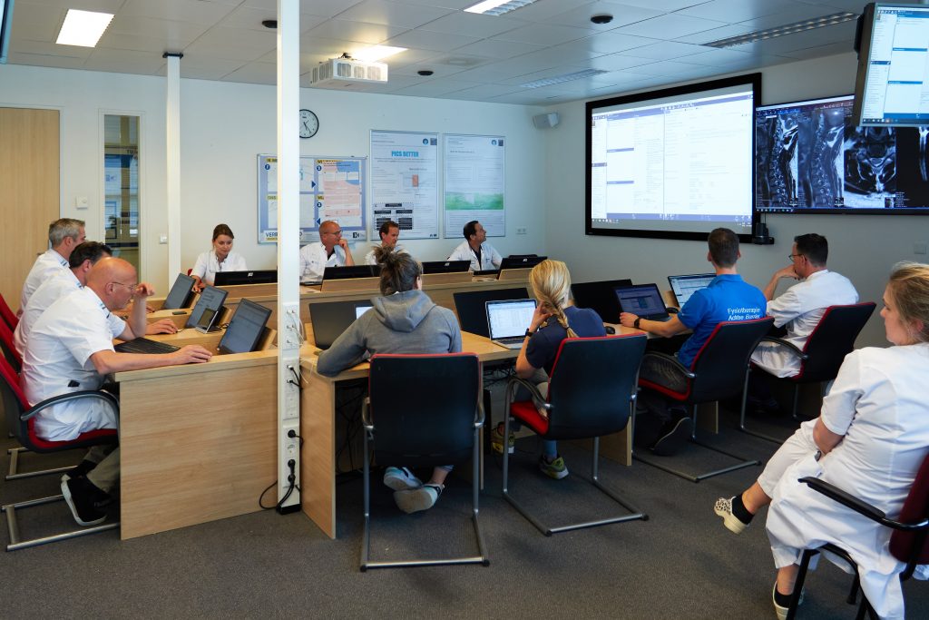 Zorgprofessionals bespreken een zorgpad voor Thoracic Outlet Syndroom rond een tafel met laptops en medische beelden op grote schermen.
