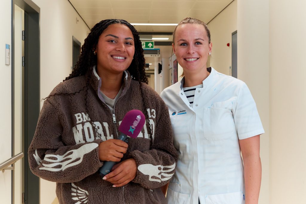 Twee vrouwen staan naast elkaar in een bende: één met microfoon, één in wit uniform. Ze delen tips over borsten checken.
