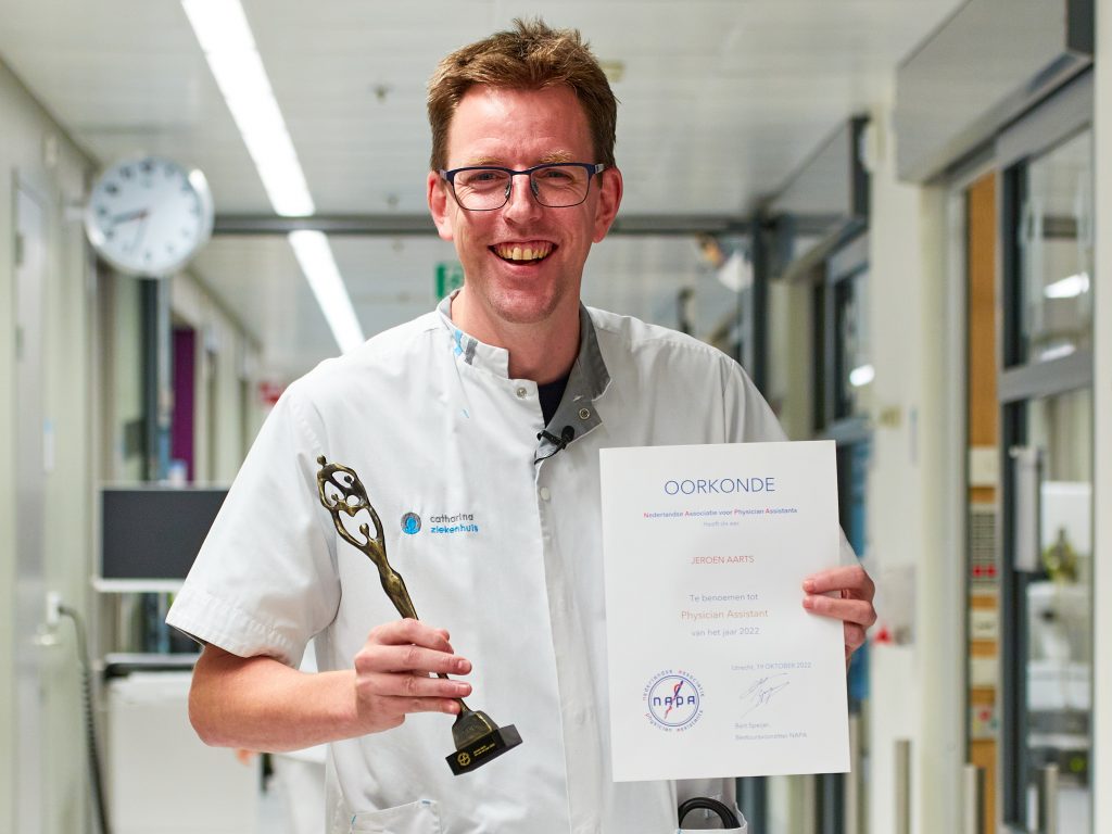 Jeroen Aarts in witte medische kleding staat in een gang met een trofee en een Oorkonde voor PA van het jaar.