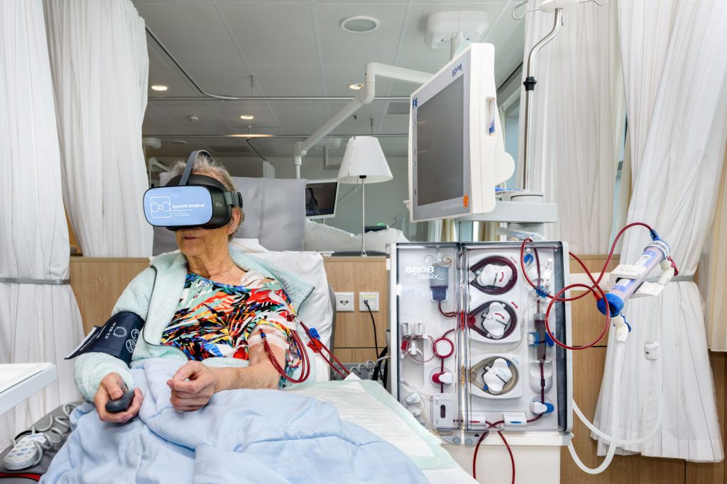 Oudere op ziekenhuisbed krijgt dialyse, draagt kleurrijk shirt en VR-bril. Omringd door medische apparatuur, Dutch Design Week.