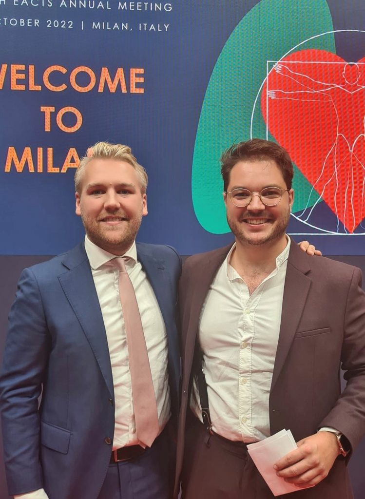 Twee mannen in pak staan naast elkaar en glimlachen. Achter hen een bord 'Welcome to Milan' met eventdetails en grafische beelden.