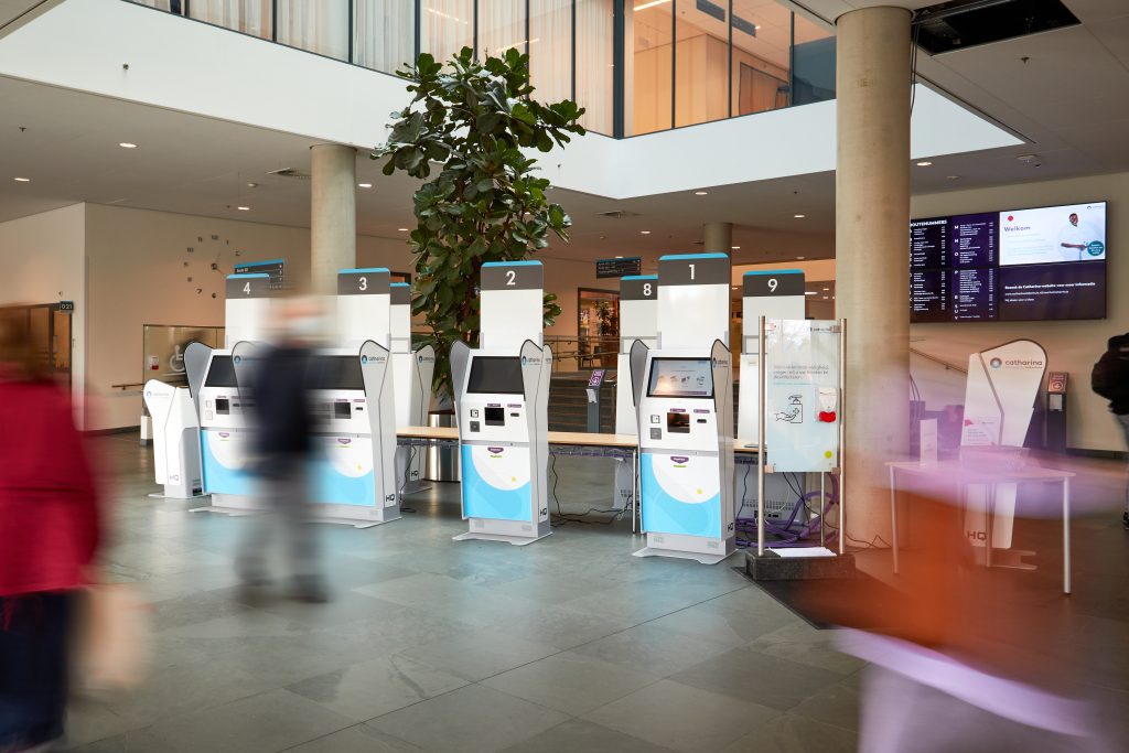 Aanmeldzuilen in een ruime lobby met grote ramen, voorbijlopende mensen, een grote plant en digitale schermen op de achtergrond.