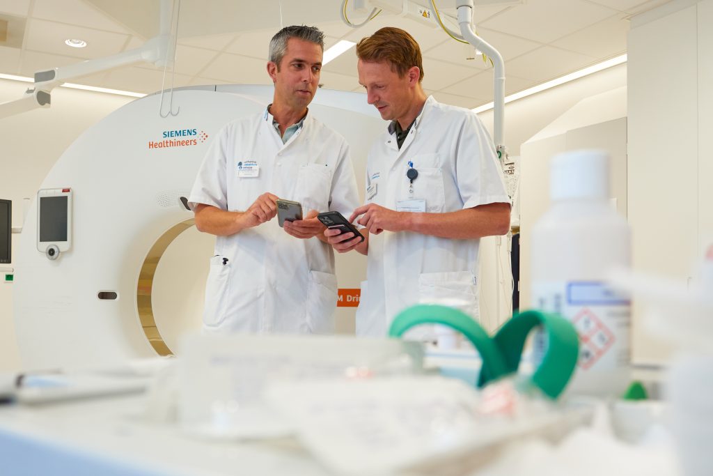 Twee artsen in witte jassen bekijken info op een mobiel bij een scanapparaat in het Catharina Ziekenhuis, met medische spullen vooraan.