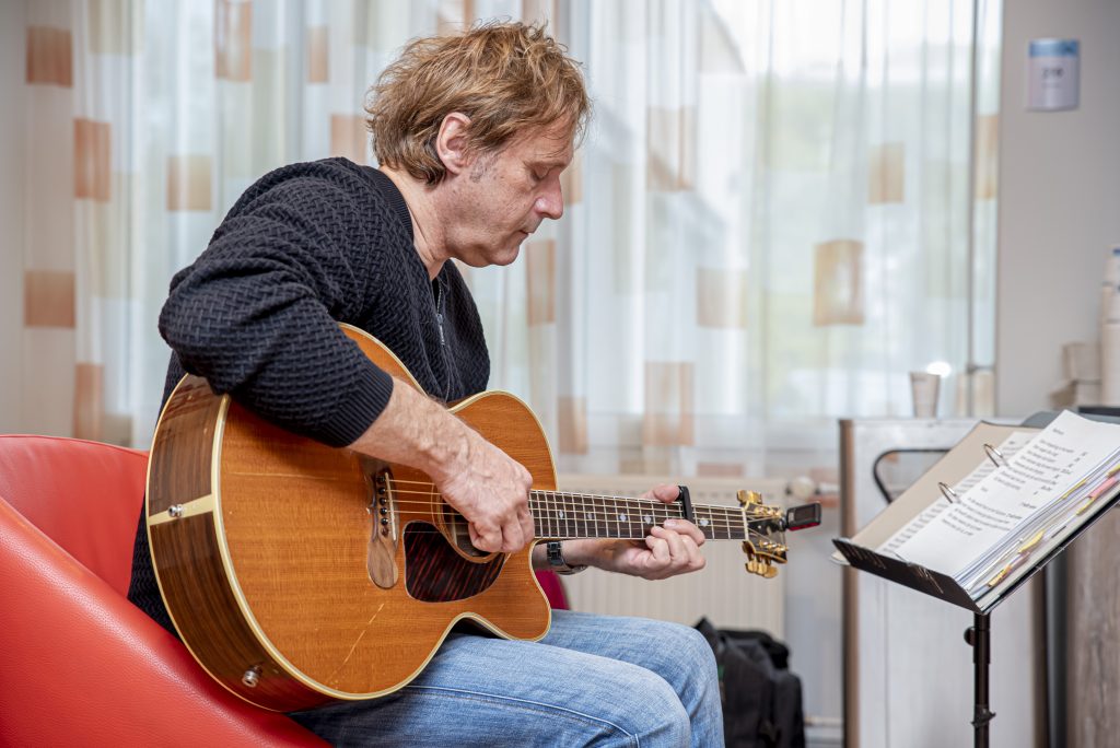 Pieter met lichtbruin haar speelt akoestische gitaar op een rode stoel, met lessenaar en gordijnen op de achtergrond.