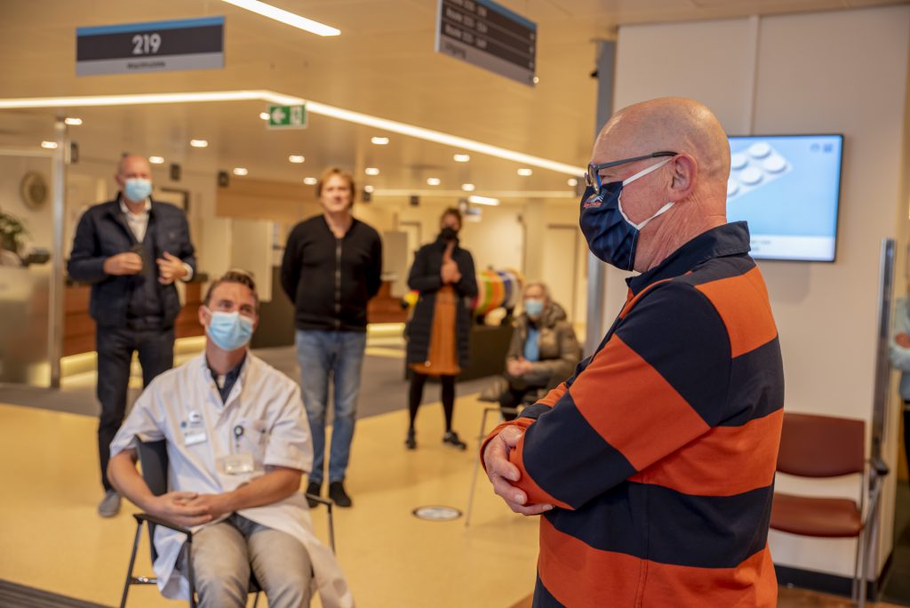 Man met gestreept shirt en mondkapje staat bij een zittende zorgmedewerker; anderen wachten in de kliniek.