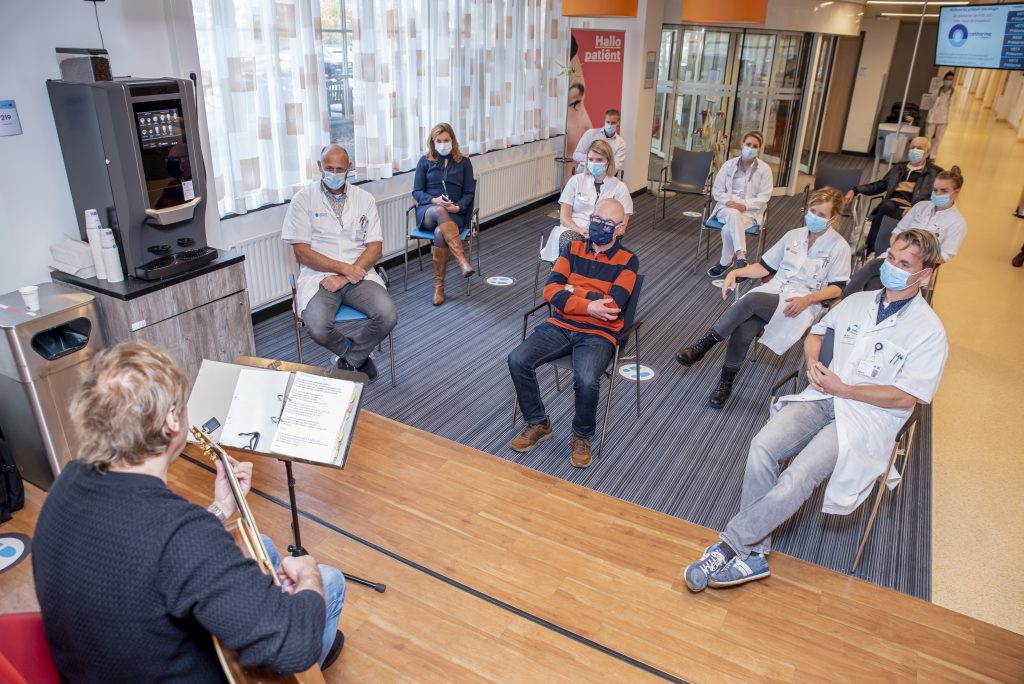 Pieter geeft een miniconcert met gitaar en zang voor mondkapdragend zorgpersoneel in een ziekenhuisgang met afstandsmarkeringen.